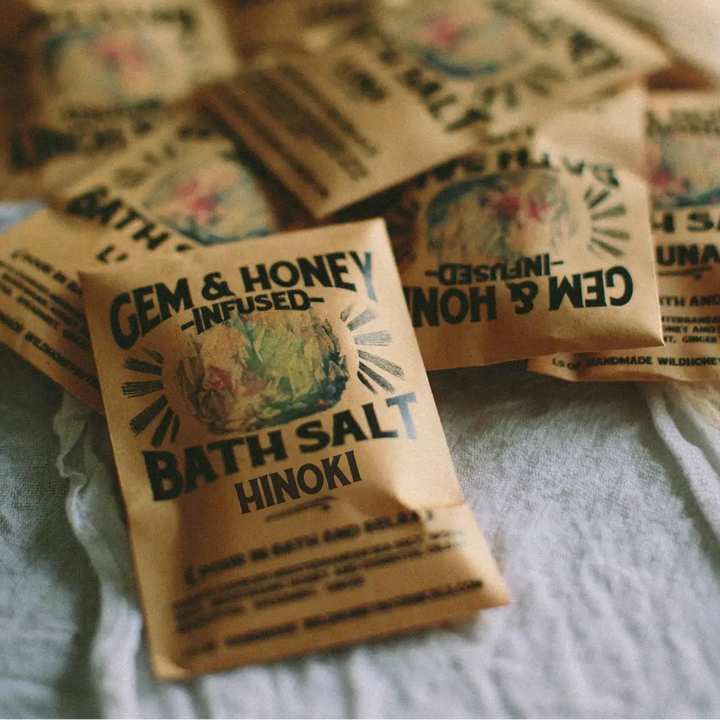 Bath salt packets labeled 'Gem & Honey Infused Bath Salt' with the text 
'HINOKI' underneath, on a textured surface. Available at Luca Essentials in Los Angeles. 