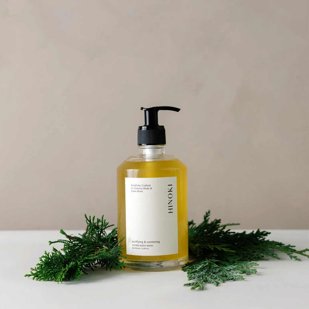 clear bottle of 'hinoki' body wash with a white label on a beige background with green leaves around the bottle. Available at Luca Essentials in Los Angeles. 