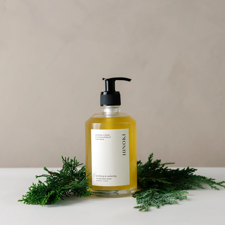 clear bottle of 'hinoki' body wash with a white label on a beige background with green leaves around the bottle. Available at Luca Essentials in Los Angeles. 
