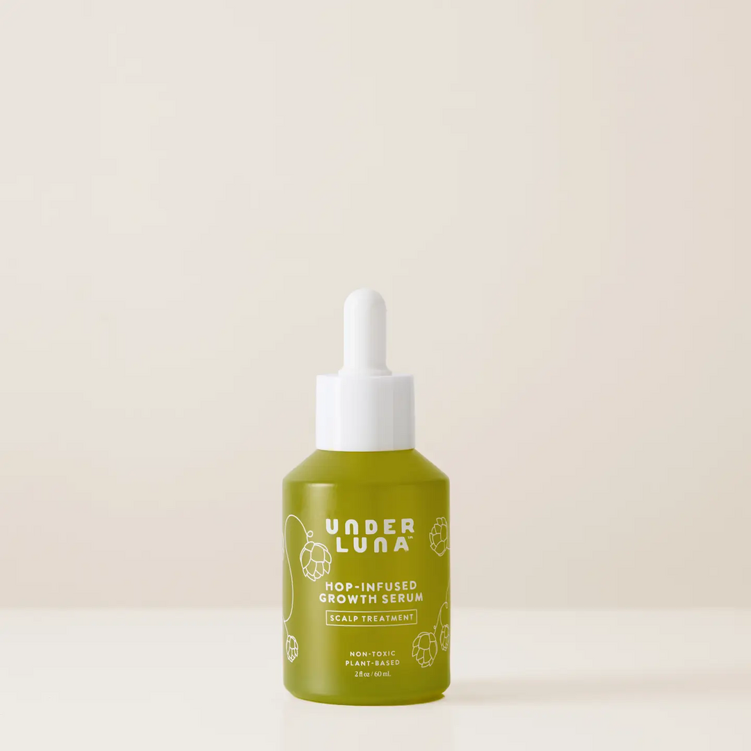 A bottle of Under Luna Hop-Infused Growth Serum with a white dropper cap and an olive green body, set against a solid cream background. Available at Luca Essentials in Los Angeles. 