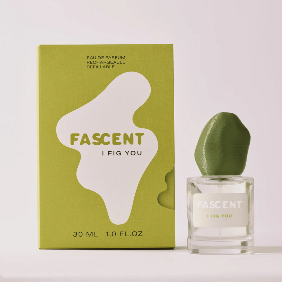 A clear glass perfume bottle with a green lid that says 'FASCENT I FIG YOU' on a light grey label and green packaging with 'Fascent' branding on a white background. Available at Luca Essentials in Los Angeles. 