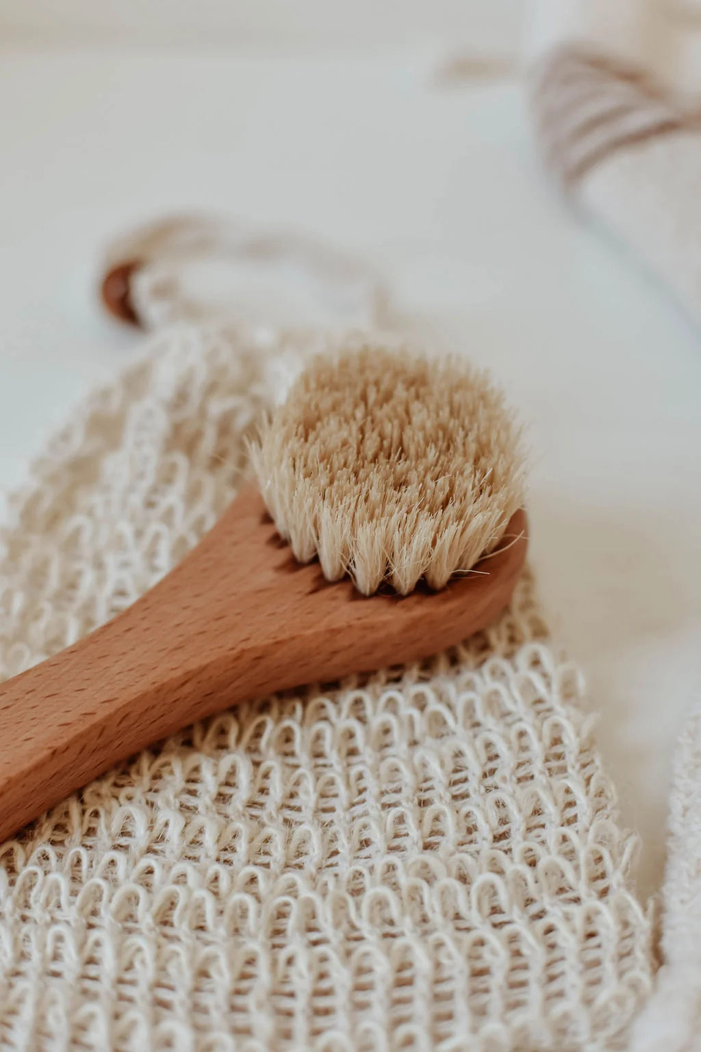 A close-up of a wooden hand brush with natural bristles resting on a textured beige washcloth. Sold at Luca Essentials in Los Angeles