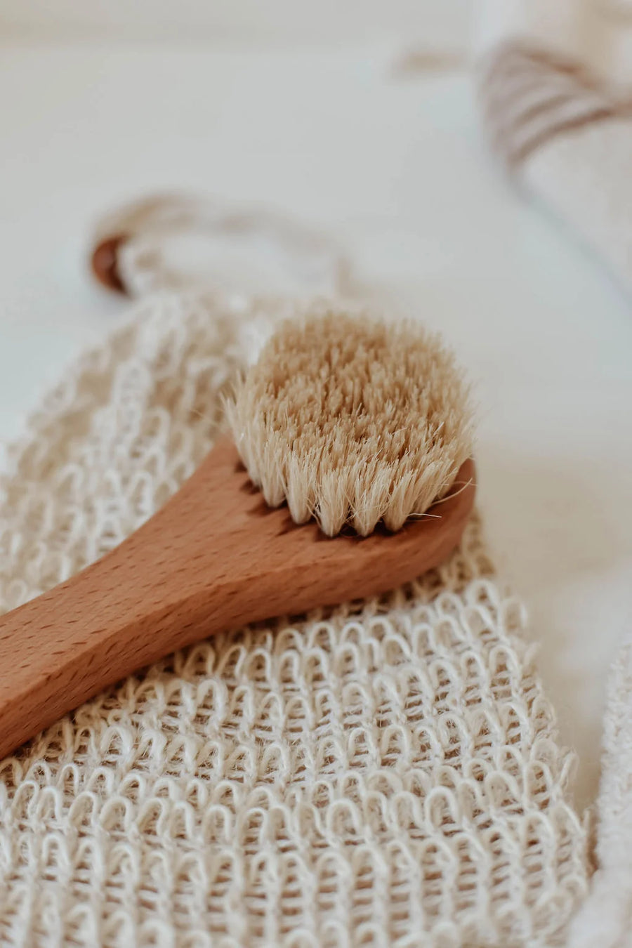 A close-up of a wooden hand brush with natural bristles resting on a textured beige washcloth. Sold at Luca Essentials in Los Angeles