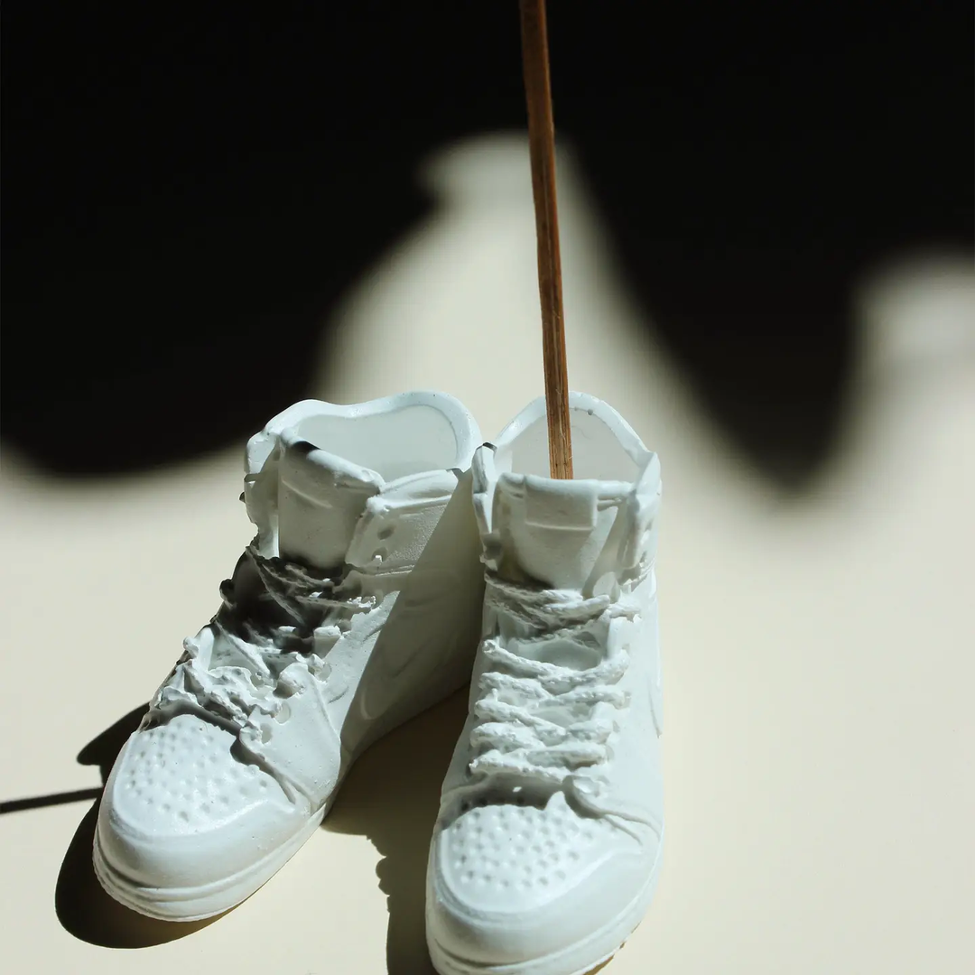 White high-top sneakers and an incense sticking out of one on a light surface with a dark background. Available at Luca Essentials in Los Angeles. 