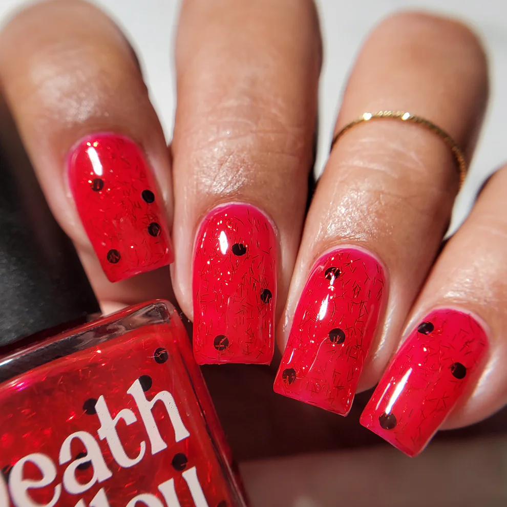A close-up of a hand with glossy red glitter nail polish featuring small black dots, shown next to a bottle labeled Death Valley Nail. Available at Luca Essentials in Los Angeles. 
