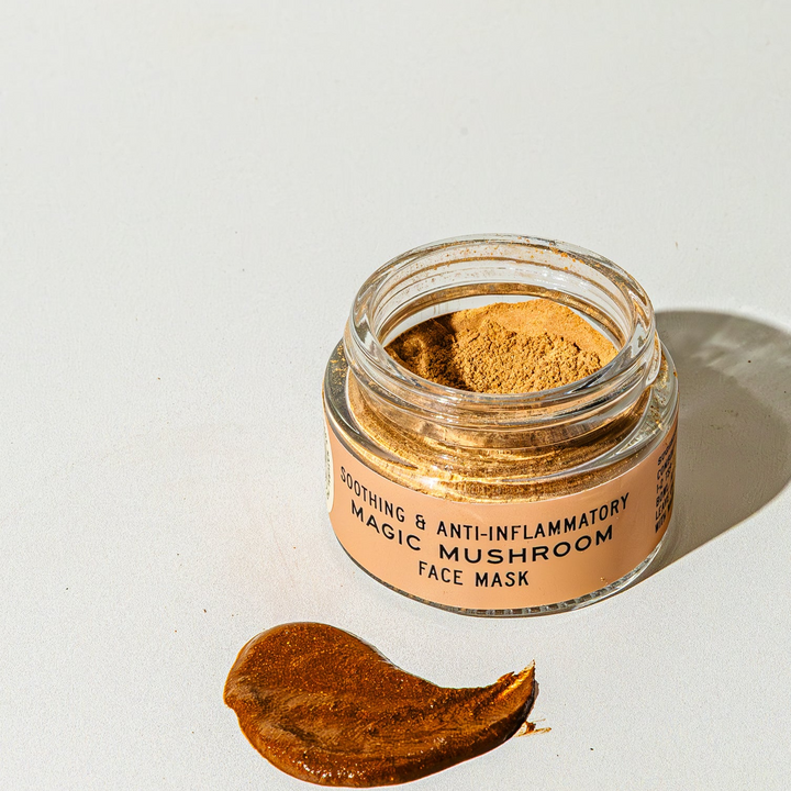 Jar of powdered magic mushroom face mask with a smear of golden brown wet mask with the words 'soothing & anti-inflammatory magic mushroom face mask' on the glass jar, on a white background. Available at Luca Essentials in Los Angeles. 