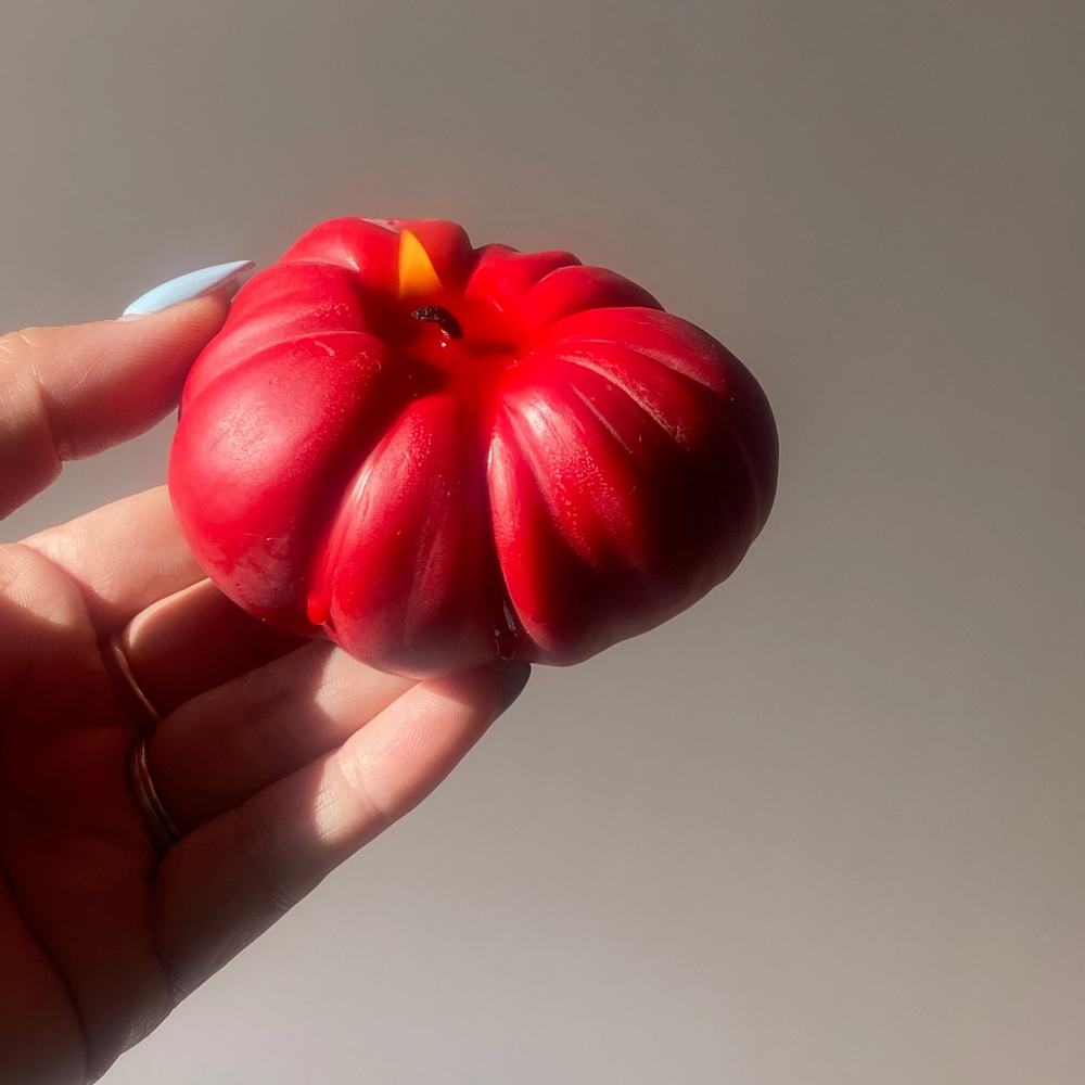 small red tomato candle with flame on wick held by a hand. 

Small but packed with personality. Our Mini Tomato Candle is a juicy little nod to peak farmer’s market season. Hand-poured in rich tomato red, it’s the tiniest member of our tomato family.

Available at Luca Essentials in Los Angeles. 