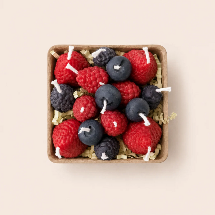 A top-down view of a square wooden tray filled with berry-shaped candles, including red raspberries, blueberries, and blackberries, arranged on a light beige background. Available at Luca Essentials in Los Angeles. 