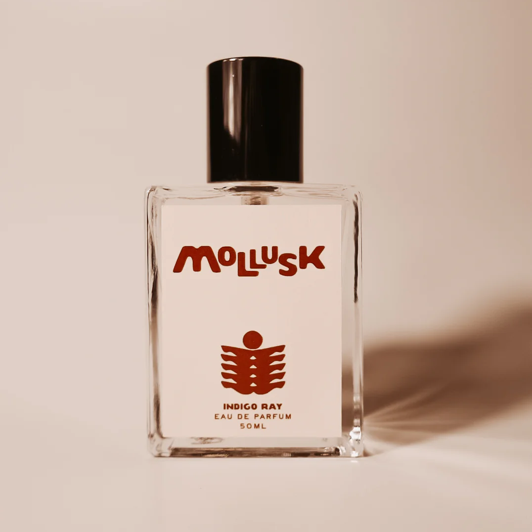 A clear glass perfume bottle with a black cap and a white label featuring the brand name 'Mollusk' in red and a red fish-like logo. The label also reads 'Indigo Ray Eau de Parfum 50ml'. Available at Luca Essentials in Los Angeles. 