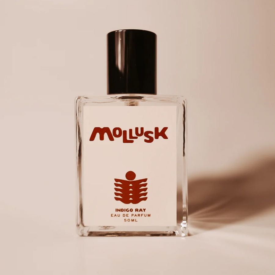A clear glass perfume bottle with a black cap and a white label featuring the brand name 'Mollusk' in red and a red fish-like logo. The label also reads 'Indigo Ray Eau de Parfum 50ml'. Available at Luca Essentials in Los Angeles. 