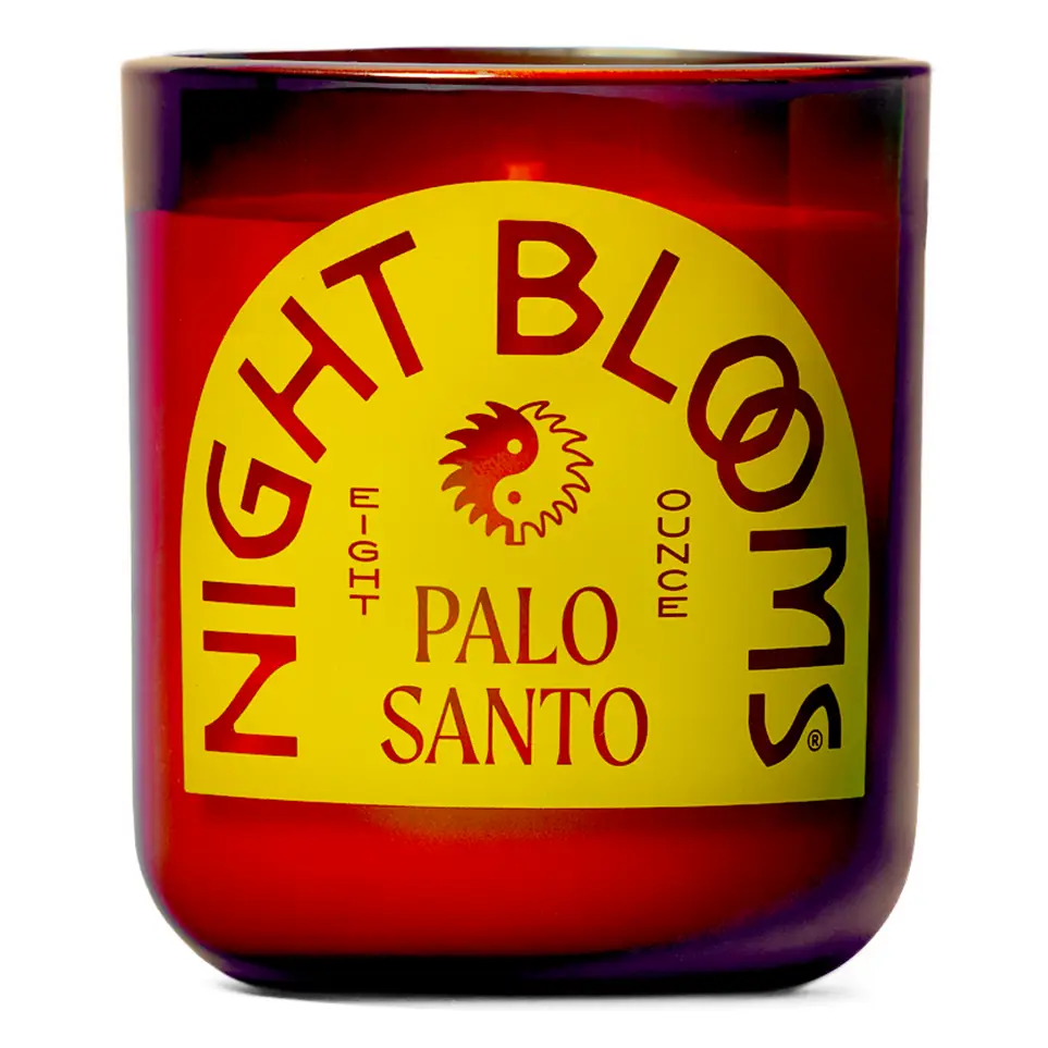 A Palo Santo candle in a translucent amber-red glass jar with a large yellow label reading 'NIGHT BLOOMS' and 'PALO SANTO' on a white background. Available at Luca Essentials in Los Angeles.