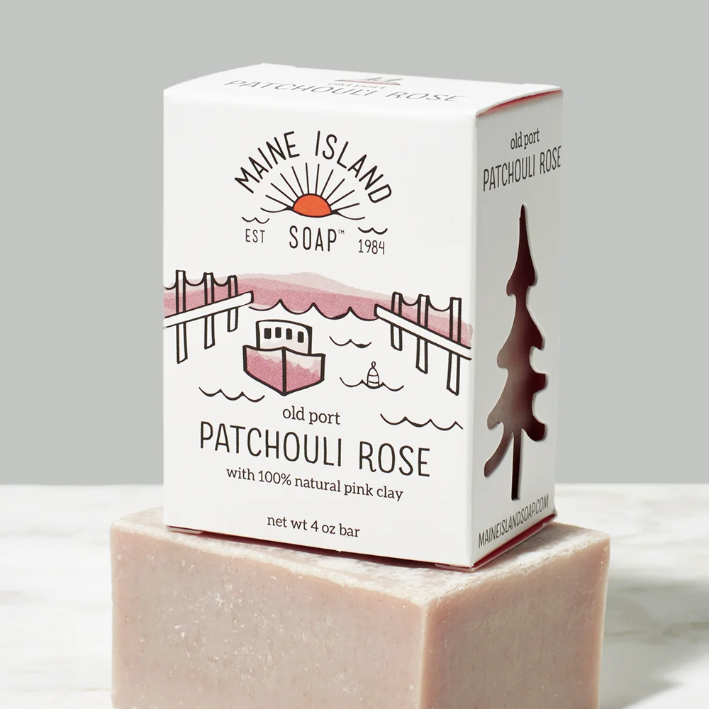 Branded soap bar in box with the logo 'Maine Island Soap' and an illustration of a boat in water between two docks. The scent 'Patchouli Rose' is located underneath the illustration. Box of bar soap is sitting on top of a beige bar soap without packaging. Available at Luca Essentials in Los Angeles. 