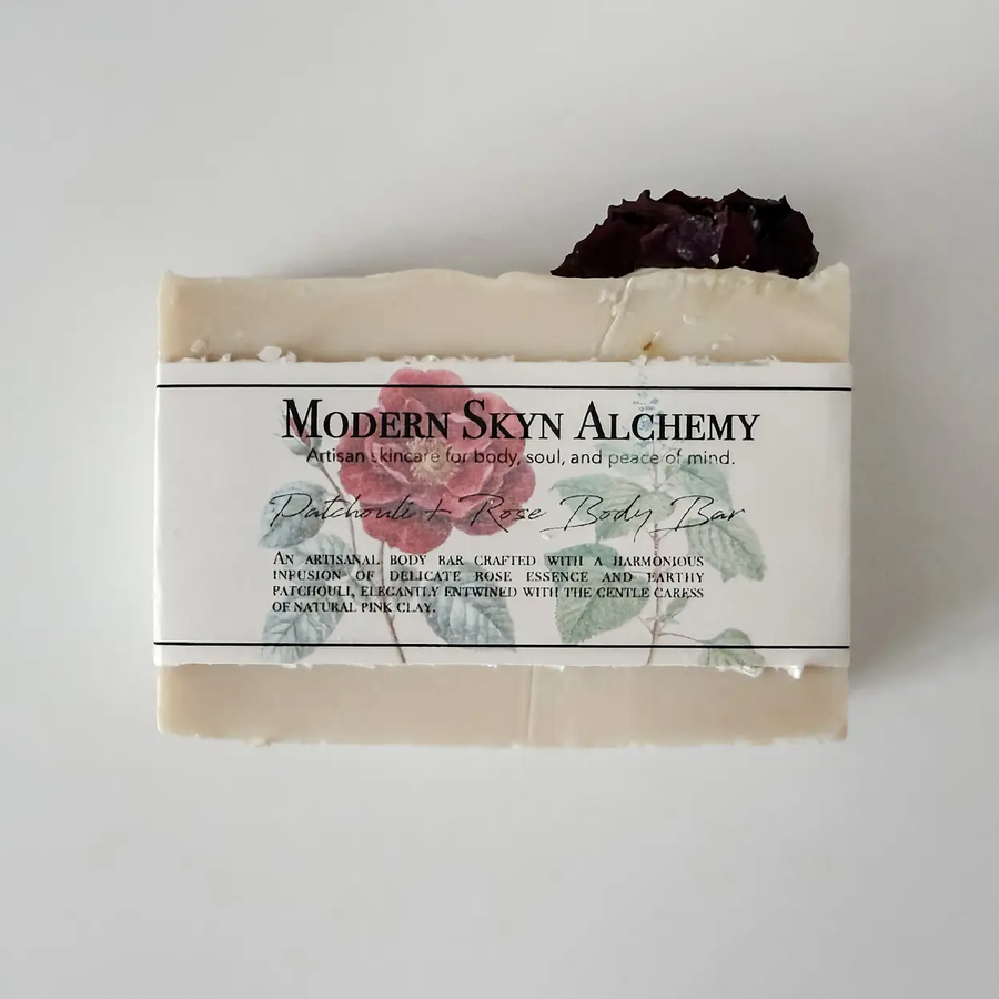 A rectangular bar of soap wrapped in a white paper label that reads 'Modern Skyn Alchemy' and 'Patchouli Rose Body Bar,' with a dark dried rose placed on top, photographed against a plain light background. Available at Luca Essentials in Los Angeles. 