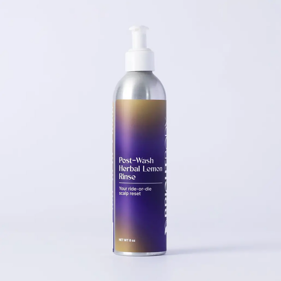 An 8 oz aluminum pump bottle of Post-Wash Herbal Lemon Rinse, featuring a purple and gold gradient label with white text, set against a clean light gray background. Available at Luca Essentials. 