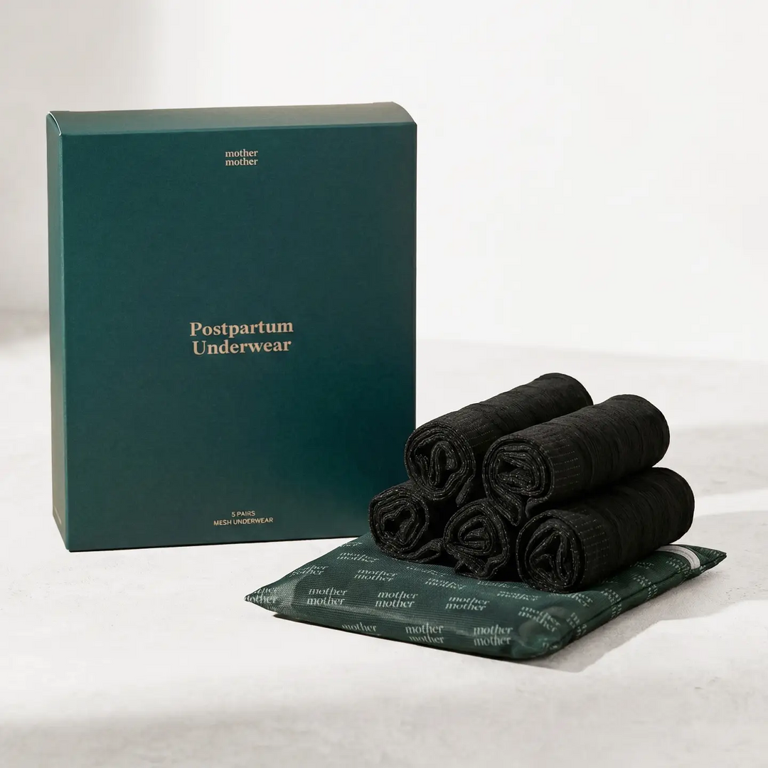 Set of 5 black postpartum underwear on top of a green mesh laundry bag with the branded 'mother mother' on it, the underwear are placed next to a green box with the product name 'postpartum underwear' on a white background. Available at Luca Essentials in Los Angeles. 