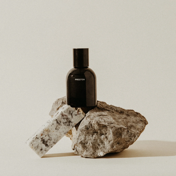 A black glass bottle with a black cap and the brand name 'PRESTON' printed in white, styled on top of stacked natural rocks against a warm off-white background. Available at Luca Essentials in Los Angeles. 