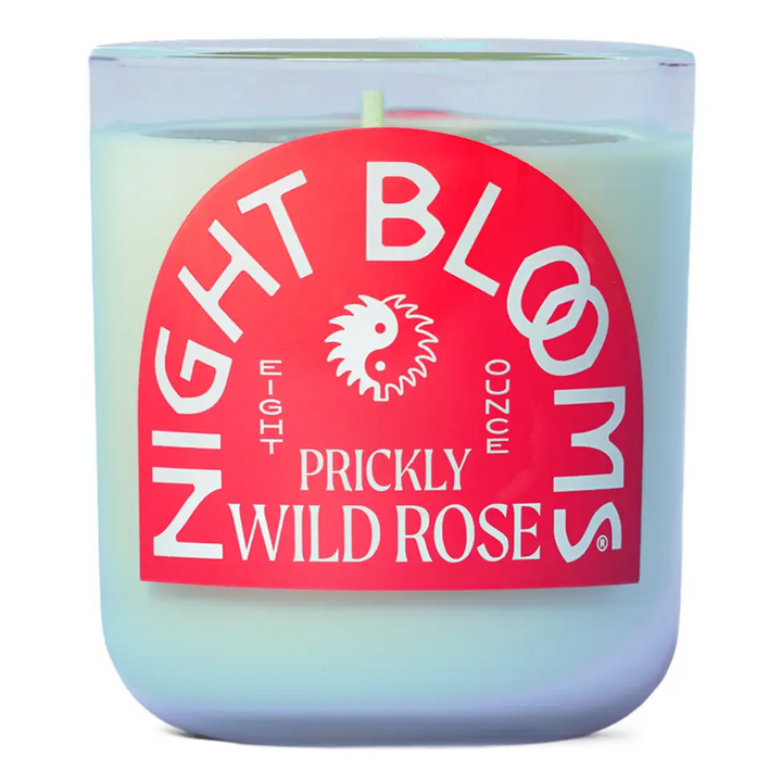 A jar candle with white wax and a large pink label reading 'NIGHT BLOOMS PRICKLY WILD ROSE' in white text, shown on a white background. Available at Luca Essentials in Los Angeles. 