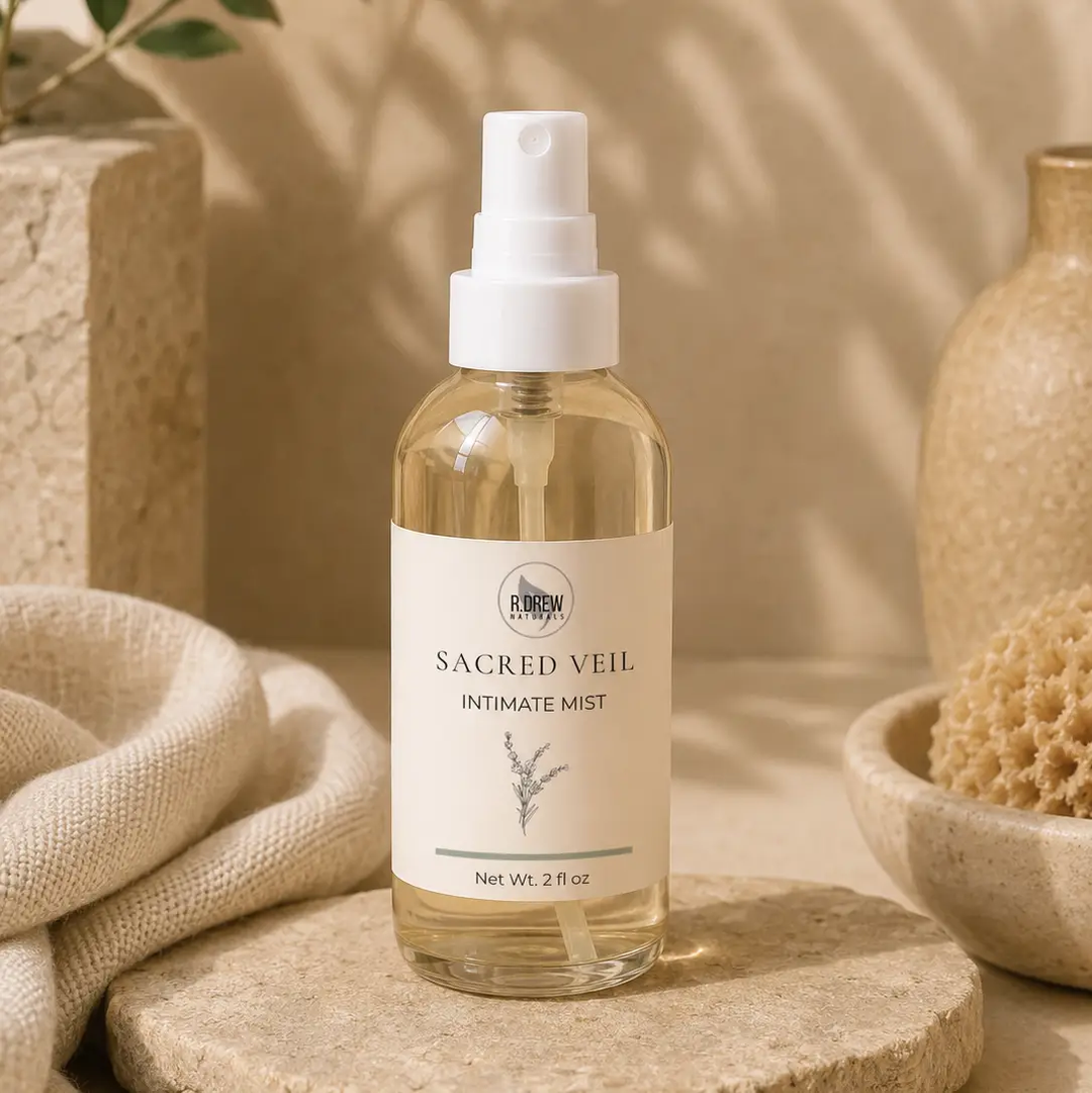 A 2 fl oz clear glass spray bottle of  Sacred Veil Intimate Mist, set on a stone slab with neutral beige props including a textured vase, a sponge, and a folded linen cloth. Available at Luca Essentials in Los Angeles. 