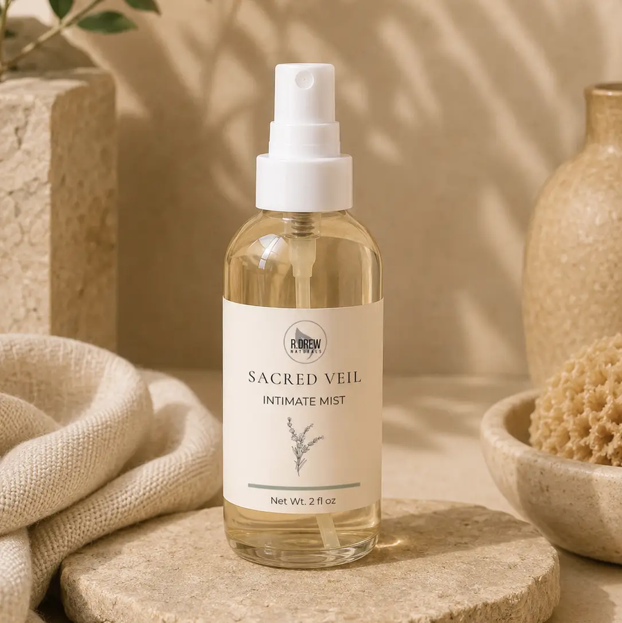 A 2 fl oz clear glass spray bottle of  Sacred Veil Intimate Mist, set on a stone slab with neutral beige props including a textured vase, a sponge, and a folded linen cloth. Available at Luca Essentials in Los Angeles. 