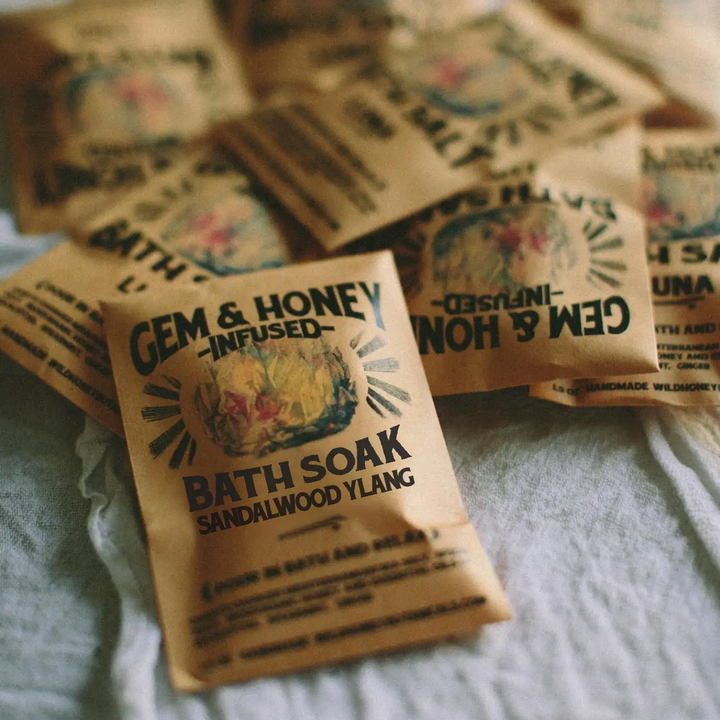 Several small brown paper packets of Gem & Honey infused bath soak, labeled Sandalwood Ylang, arranged on a light fabric surface. Available at Luca Essentials in Los Angles. 