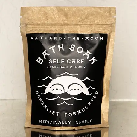 A brown kraft paper resealable bag with a black and white label on the front with the words 'BATH SOAK SELF CARE' and an illustration of a moon with eyes in waves. The bag is placed on top of a mirrored surface with a white wall in the background. Available at Luca Essentials in Los Angeles. 