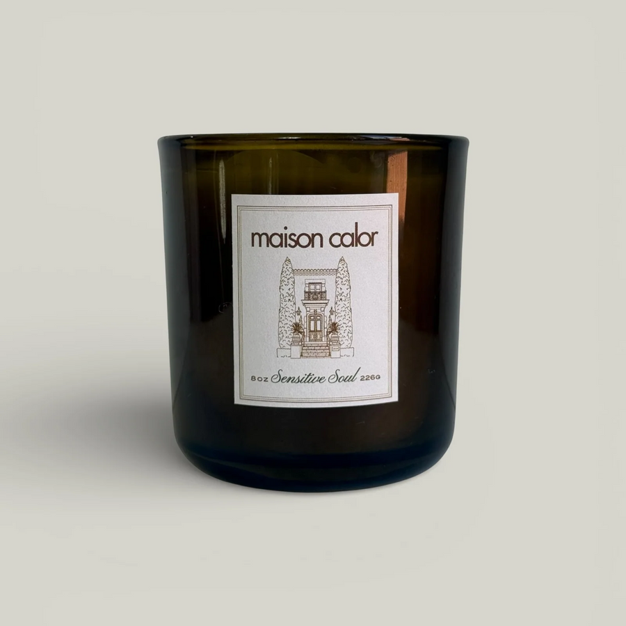 A dark amber glass jar candle with a white rectangular label reading 'maison calor' and a small architectural illustration, photographed on a plain light warm-gray background. Available at Luca Essentials in Los Angeles. 