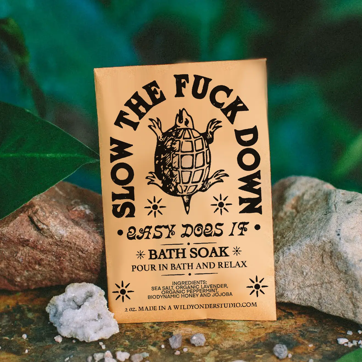 Bath soak packaging with turtle illustration and text that says 'SLOW THE FUCK DOWN EASY DOES IT BATH SOAK' propped up on two rocks on a natural background. Available at Luca Essentials in Los Angeles. 