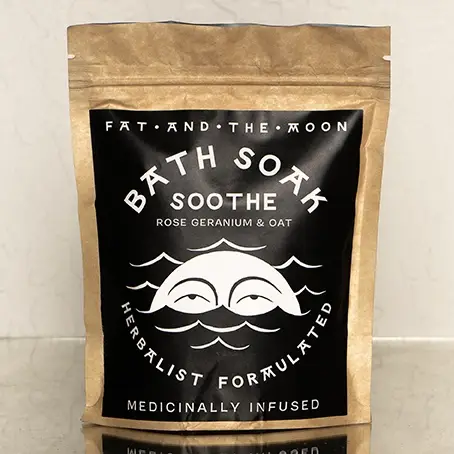 A brown kraft paper resealable bag with a black and white label on the front with the words 'BATH SOAK SOOTHE' and an illustration of a moon with eyes in waves. The bag is placed on top of a mirrored surface with a white wall in the background. Available at Luca Essentials in Los Angeles. 
