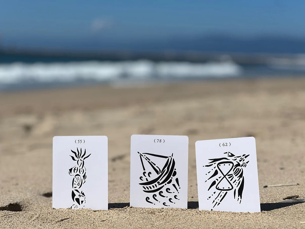 three white and black oracle cards in sand at the beach with waves in the background. Available at Luca Essentials in Los Angeles. 