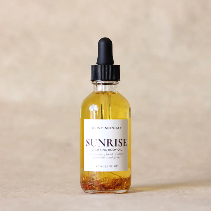Glass bottle with black dropper lid with the word "sunrise body oil' on a white sticker on the bottle on a beige background. Available at Luca Essentials in Los Angeles 