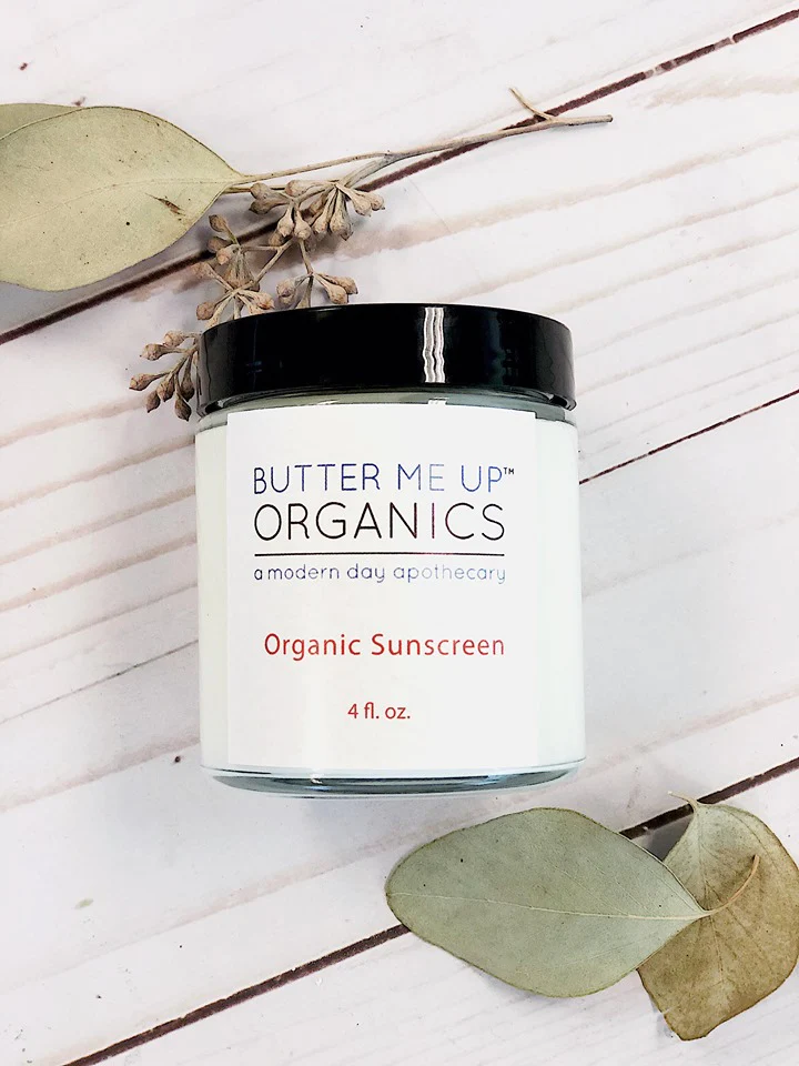A clear jar with the label that reads "Butter Me Up Organics(tm) Organic Sunscreen. A modern day apothercary" with a black lid, containing a white cream. The label includes the brand name and product details. Butter Me Up Organics is available at Luca Essentials.