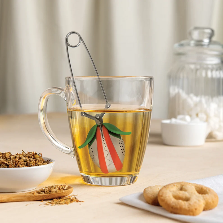 Clear mug with tea and a venus fly trap shaped tea infuser, surrounded by tea leaves and cookies on a wooden surface. Available at Luca Essentials in Los Angeles. 