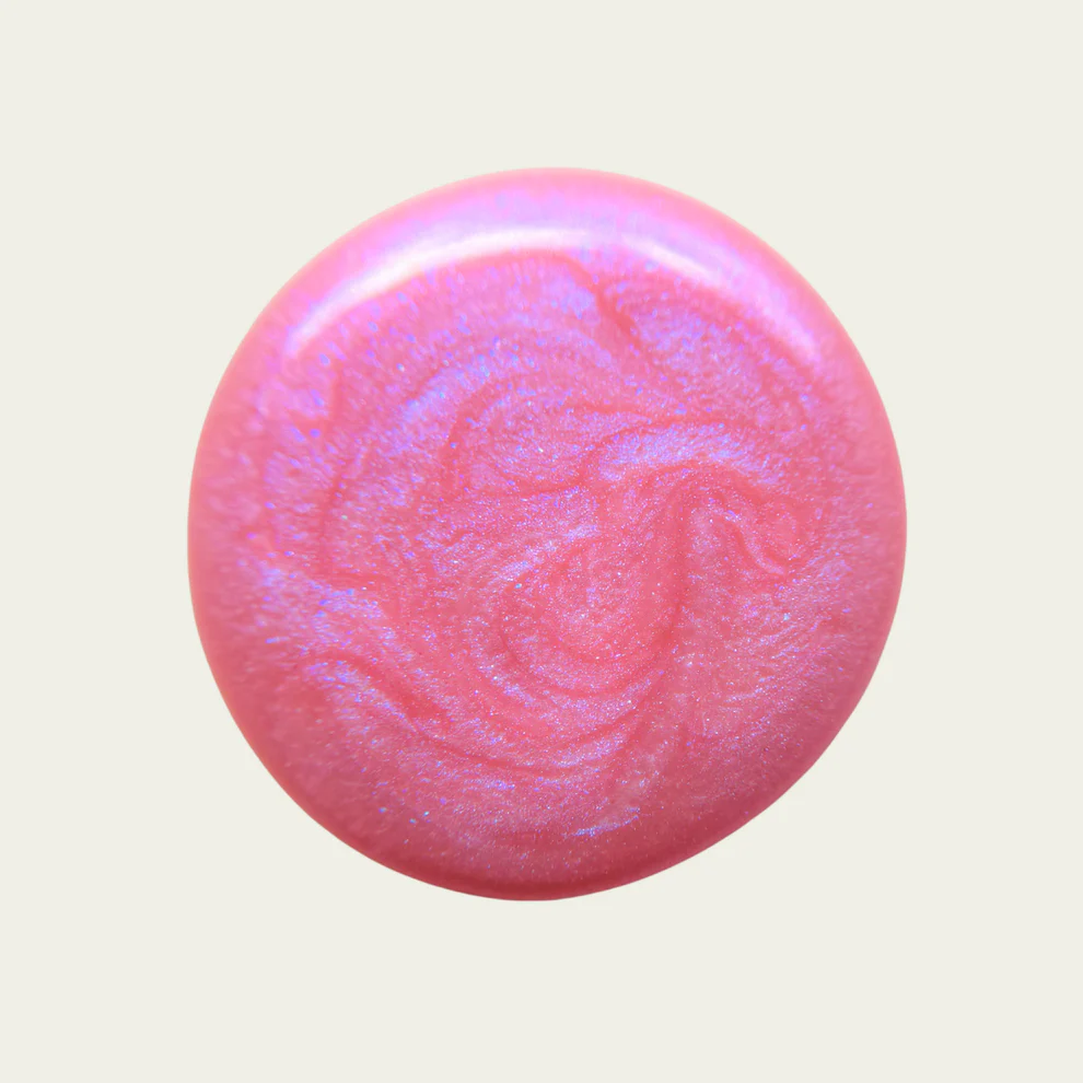 A round, glossy pink nail polish swatch with a subtle iridescent shimmer, set against a solid light beige background. Available at Luca Essentials in Los Angeles. 