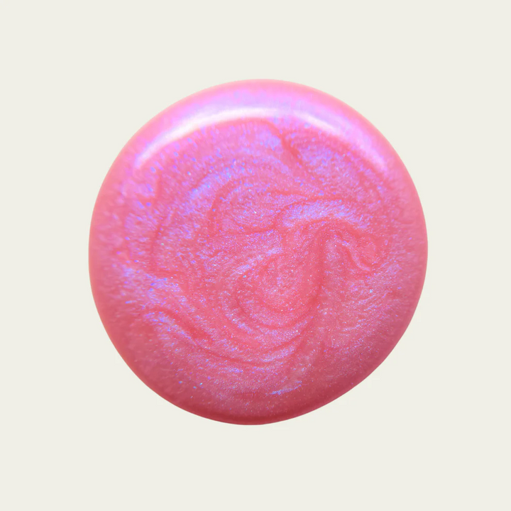 A round, glossy pink nail polish swatch with a subtle iridescent shimmer, set against a solid light beige background. Available at Luca Essentials in Los Angeles. 