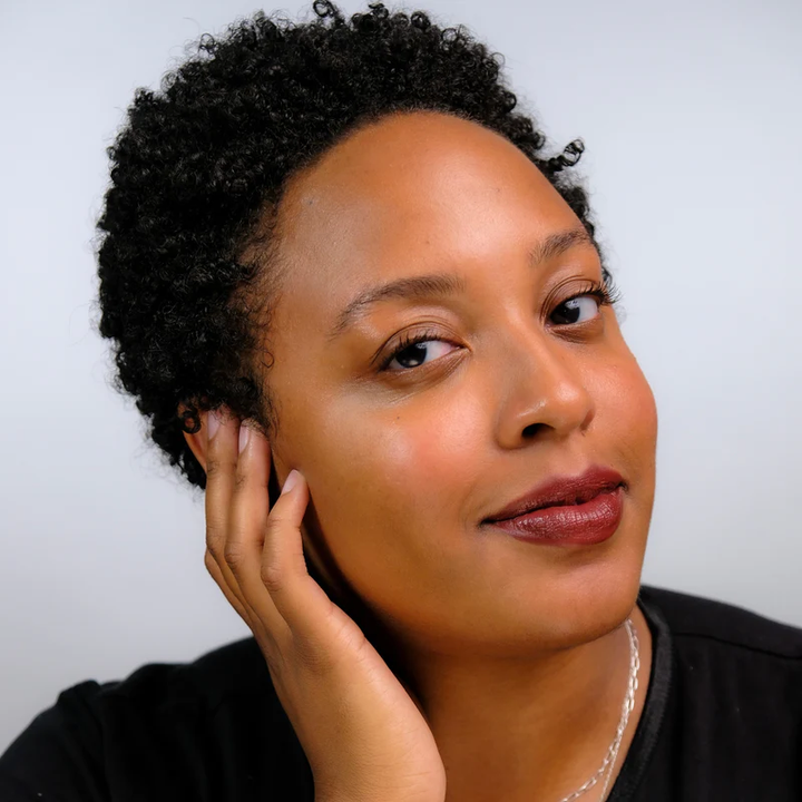 Topaz - Medium skin toned woman with short curly hair touching her face against a plain background. Available at Luca Essentials in Los Angeles. 