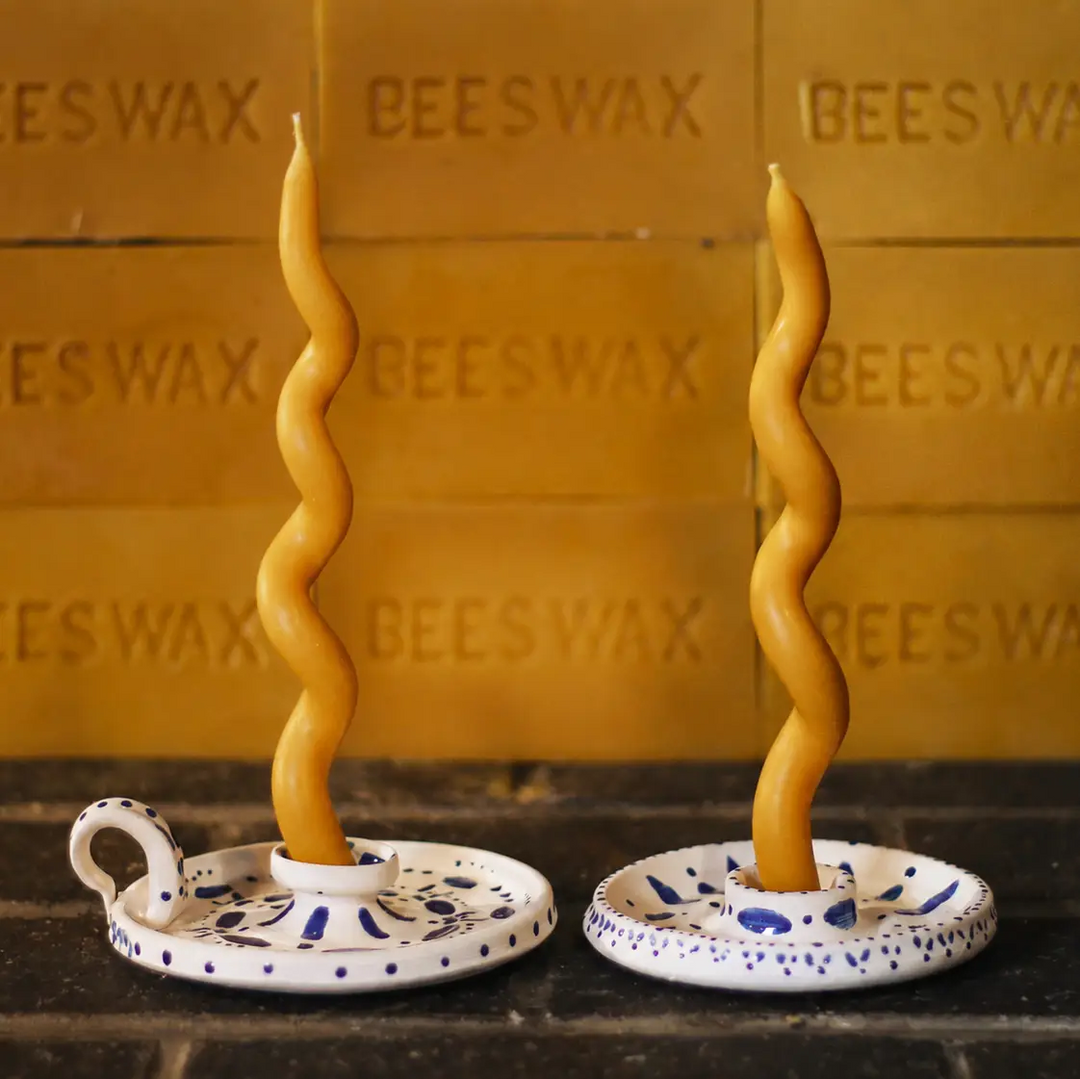 Two twisted beeswax taper candles in white and blue speckled ceramic holders, set against a warm honey-colored beeswax block background. Available at Luca Essentials in Los Angeles. 