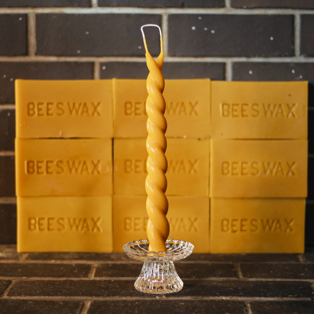 A twisted beeswax taper candle in a clear glass pedestal holder, set against a background of stacked yellow beeswax bars embossed with the word BEESWAX. Available at Luca Essentials in Los Angeles. 
