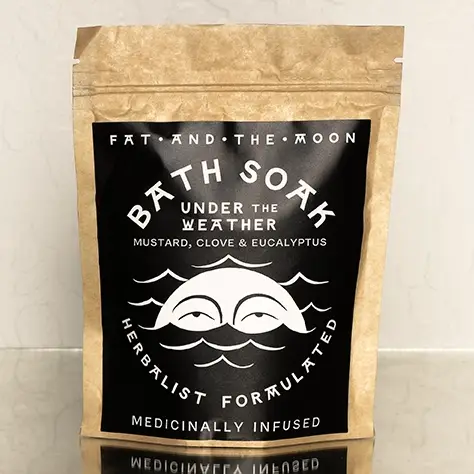A brown kraft paper resealable bag with a black and white label on the front with the words 'BATH SOAK UNDER THE WEATHER' and an image of half a sun with eyes inside of waves on a mirrored ground and white background. Available at Luca Essentials in Los Angeles. 