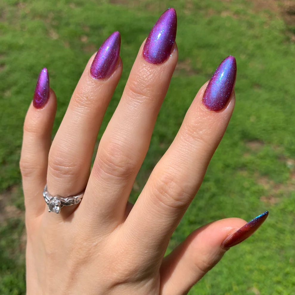 A close-up of a hand with long, stiletto-shaped nails painted in a metallic purple and blue iridescent finish, with a silver ring on the ring finger, set against a blurred green grass background. Available at Luca Essentials in Los Angeles. 