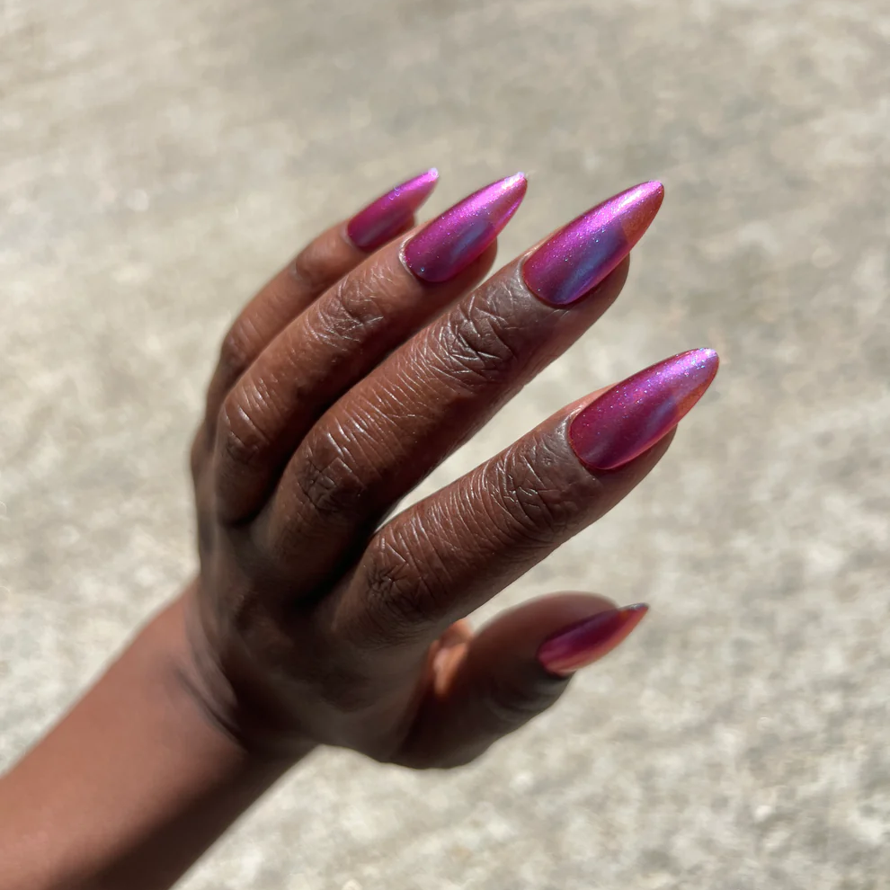 A close-up of a hand with long, almond-shaped nails painted in a metallic magenta and purple iridescent nail polish, set against a blurred concrete background. Available at Luca Essentials in Los Angeles. 