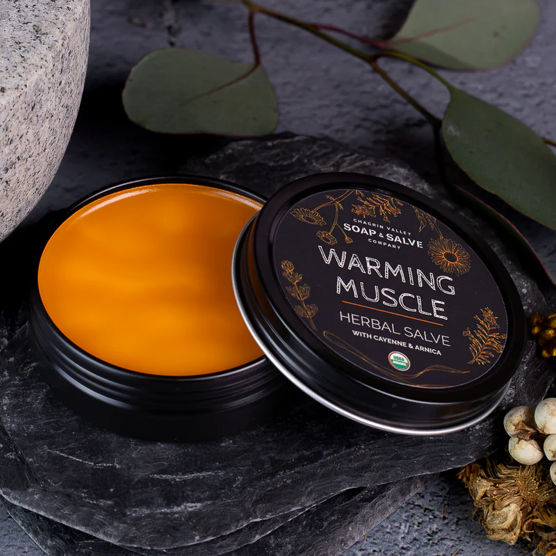 Black tin and lid propped up against the other tin with orange ointment inside on a black background with eucalyptus leaves in the background. Available at Luca Essentials in Los Angeles. 