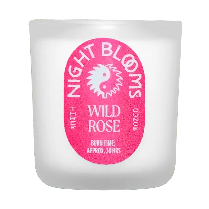A frosted glass candle jar with a prominent pink oval label reading 'NIGHT BLOOMS WILD ROSE' and 'BURN TIME: APPROX. 20 HRS'. Available at Luca Essentials in Los Angeles. 