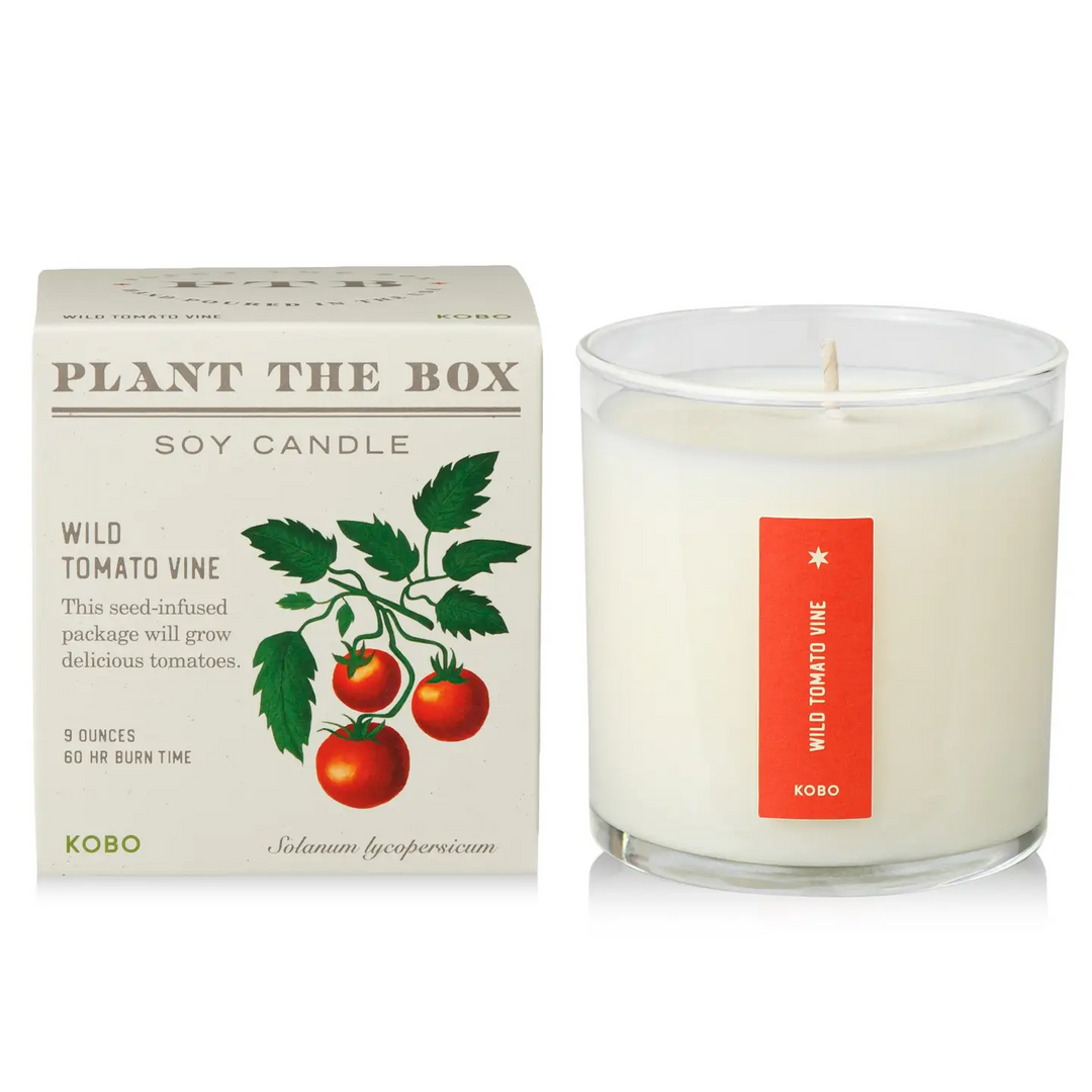 A glass candle with a red sticker down the middle with the words 'wild tomato vine' next to it's packaging with the product name 'plant the box soy candle' on a white background. Available at Luca Essentials in Los Angeles. 